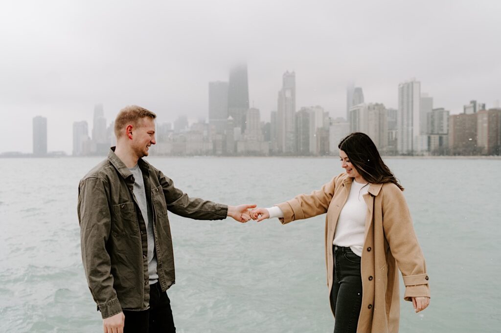 A man and woman smile at one another while holding hands in front of Lake Michigan and the Chicago skyline during their Fall Engagement Session at North Avenue Beach