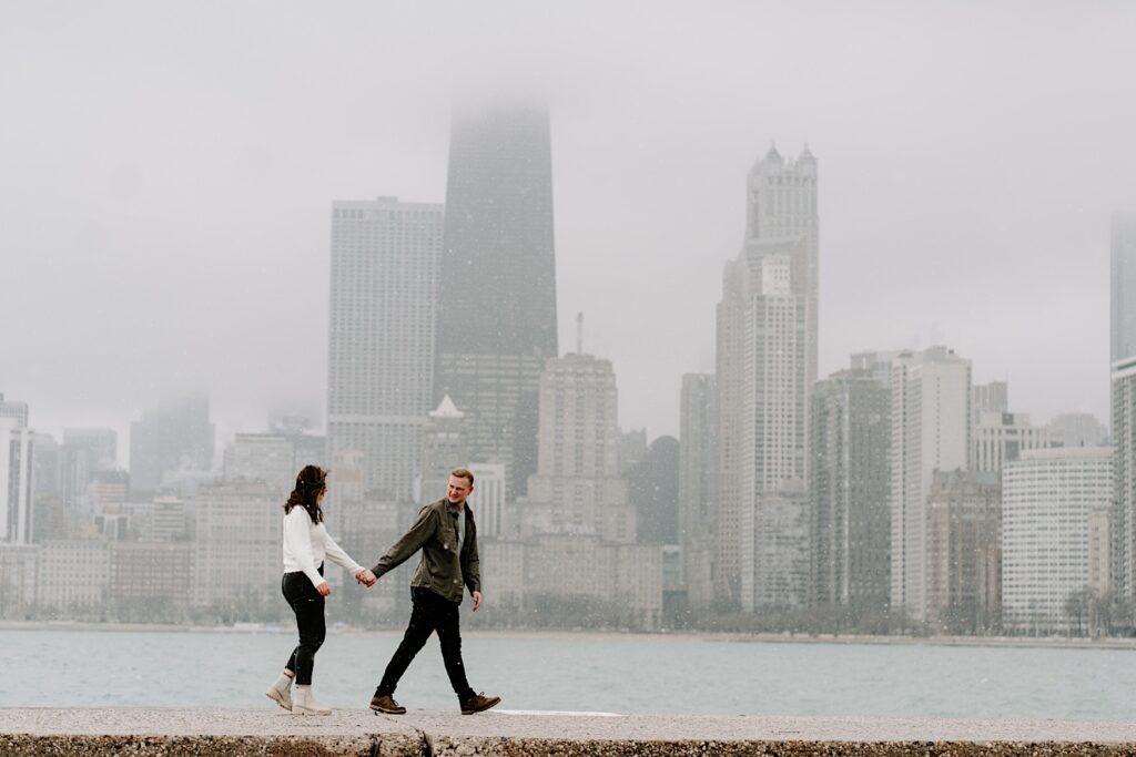 A man and woman walk hand in hand with one another as the man looks back at her with Lake Michigan and the Chicago skyline behind them during their Fall Engagement Session at North Avenue Beach