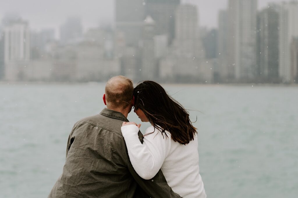 A man and woman sit next to one another and face away from the camera looking towards Lake Michigan and the Chicago skyline during their Fall Engagement Session at North Avenue Beach
