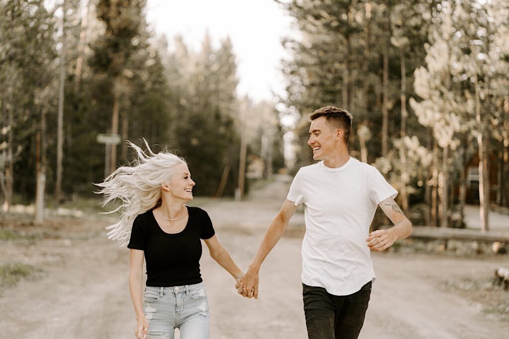 A man and woman run hand in hand and smile at one another down a road in the middle of the Busse Woods Forest Preserve during their engagement session