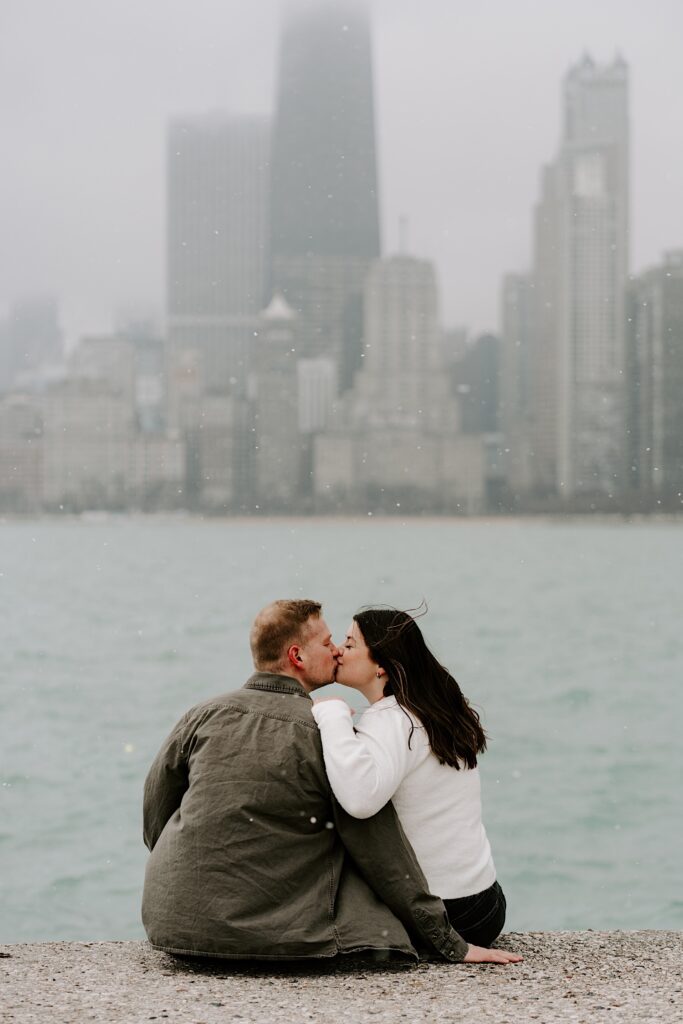 A man and woman sit next to one another on the shore of Lake Michigan and kiss one another with the Chicago skyline behind them