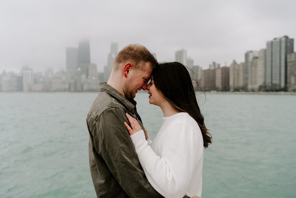 A man and woman embrace one another and smile at each other with their noses touching in front of Lake Michigan and the Chicago skyline during their Fall Engagement Session at North Avenue Beach