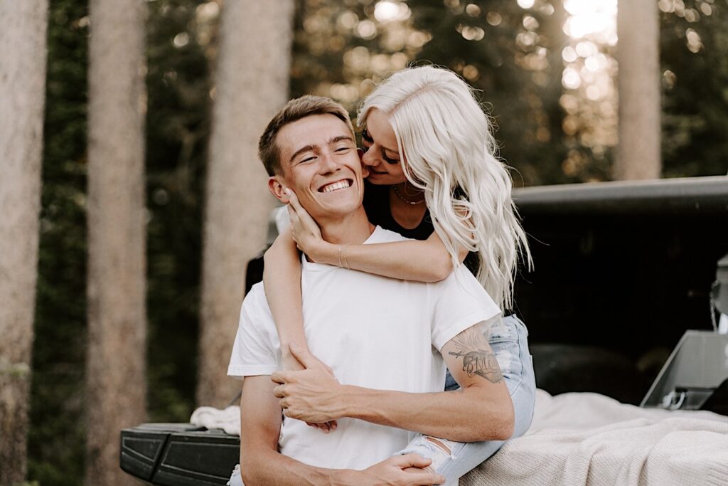 A man smiles as a woman behind him bites his ear as the two sit in the back of a truck bed together during their engagement session at Busse Woods Forest Preserve