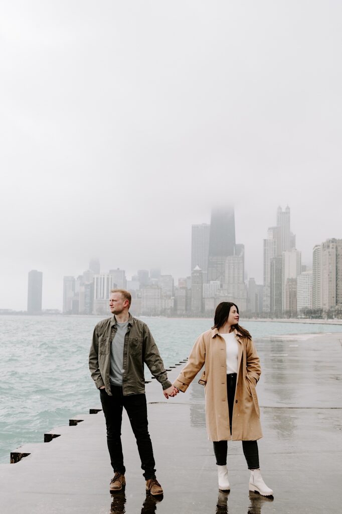 A man and woman hold hands and face the camera while looking in opposite directions with Lake Michigan and the Chicago skyline behind them