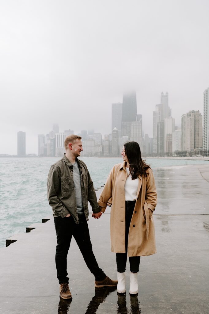 A man and woman hold hands while smiling at one another with Lake Michigan and the Chicago skyline behind them