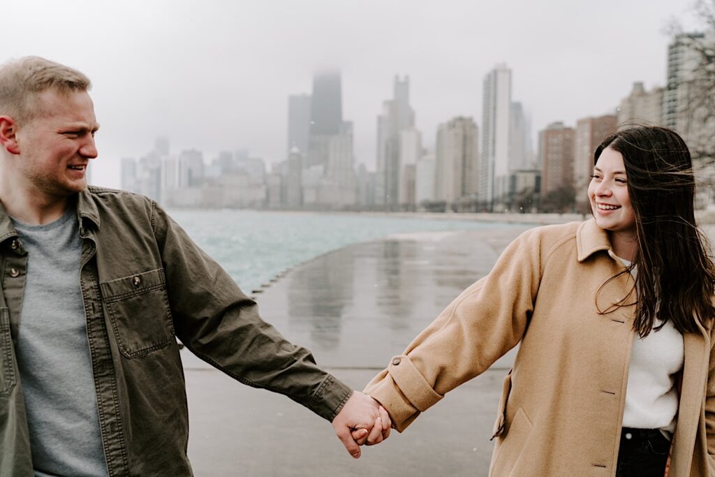 A man and woman hold hands and smile at one another with Lake Michigan and the Chicago skyline behind them during their Fall Engagement Session at North Avenue Beach