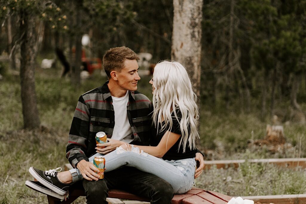 A man and woman sit next to one another looking and smiling at each other with a beer in either of their hands during their engagement session in the middle of Busse Woods Forest Preserve