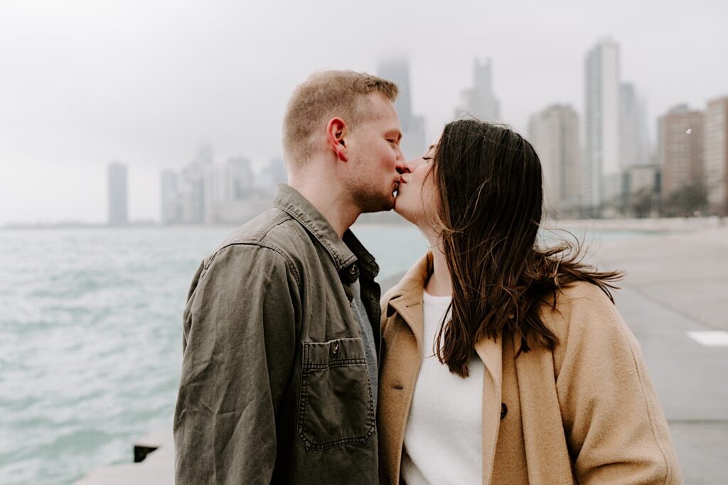 A man and woman kiss one another with their eyes closed while standing in front of Lake Michigan and the Chicago skyline during their Fall Engagement Session at North Avenue Beach