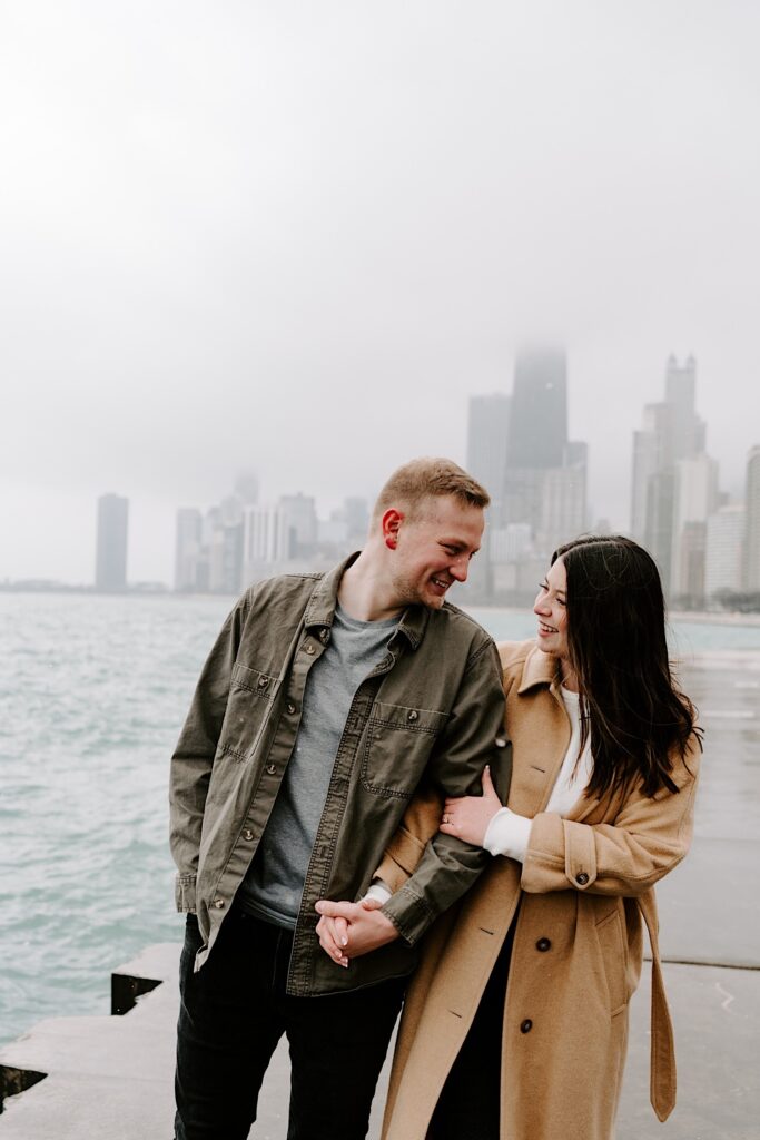A man and woman walk hand in hand next to one another while smiling at each other with the Chicago skyline and Lake Michigan behind them