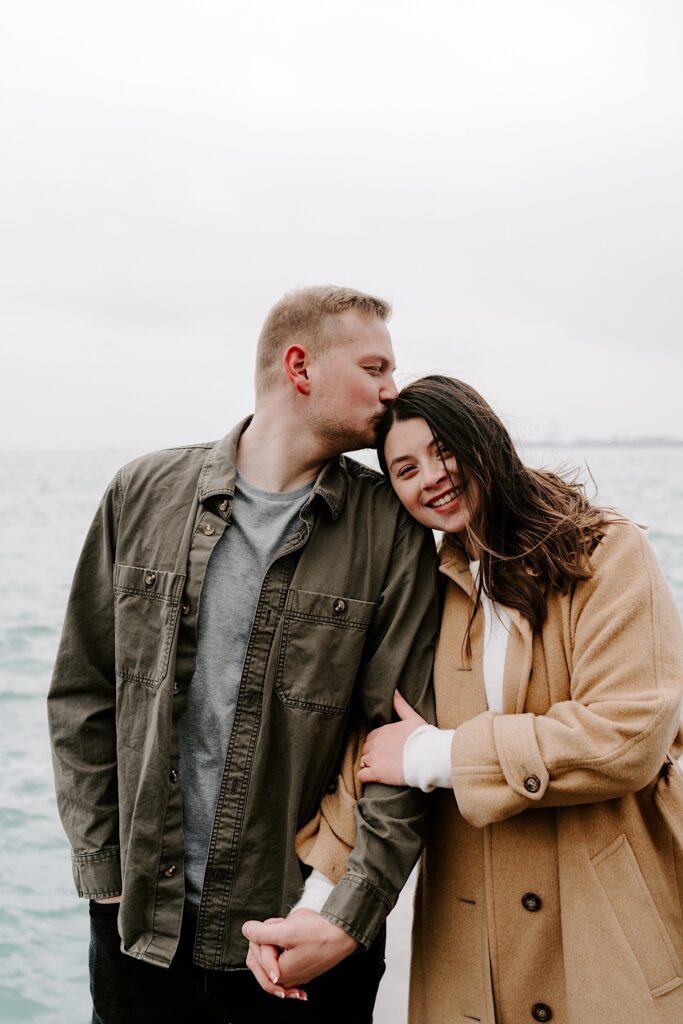 A woman smiles at the camera as a man holds her hand and kisses her on the head in front of Lake Michigan