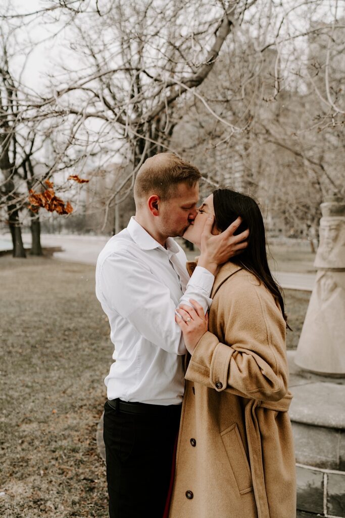 A man and woman kiss one another while in a park in Chicago with bare trees and buildings behind them