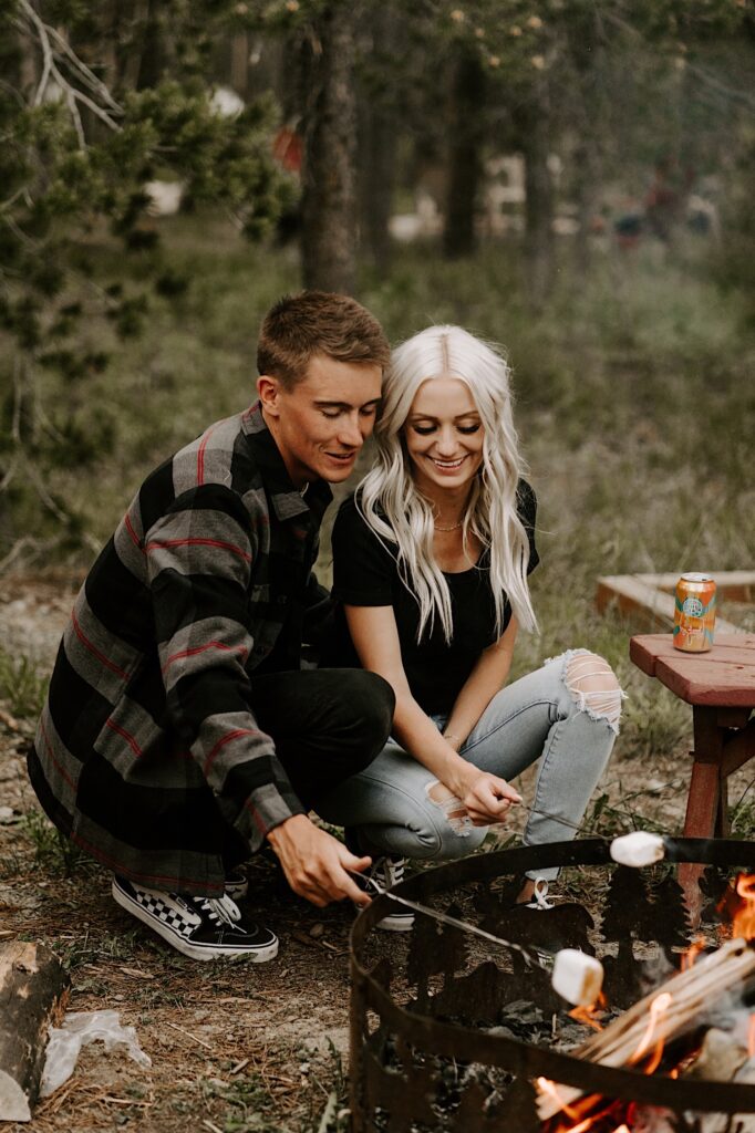 A man and woman crouch next to one another and roast marshmallows in the middle of a forest