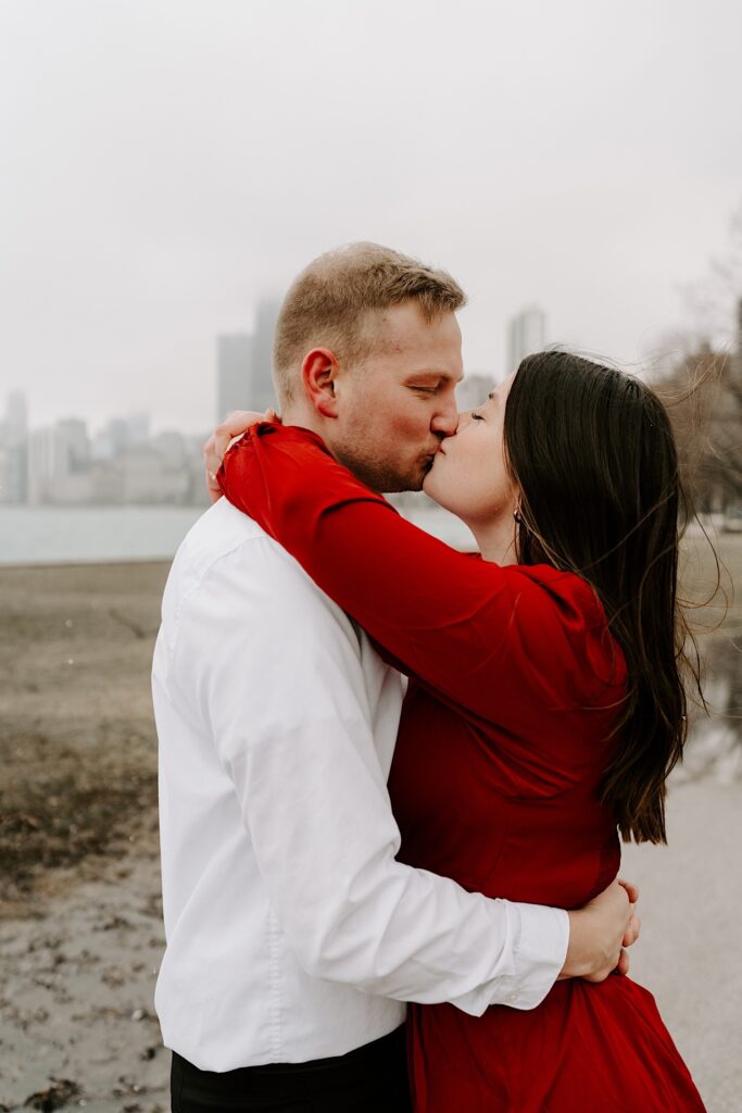 A man and woman kiss one another and embrace in a park in Chicago with the city skyline and Lake Michigan behind them