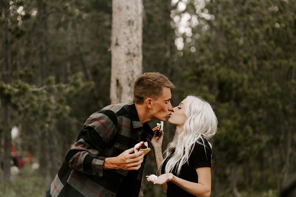 A man and woman kiss one another while holding smores in the middle of Busse Woods Forest Preserve during their engagement session