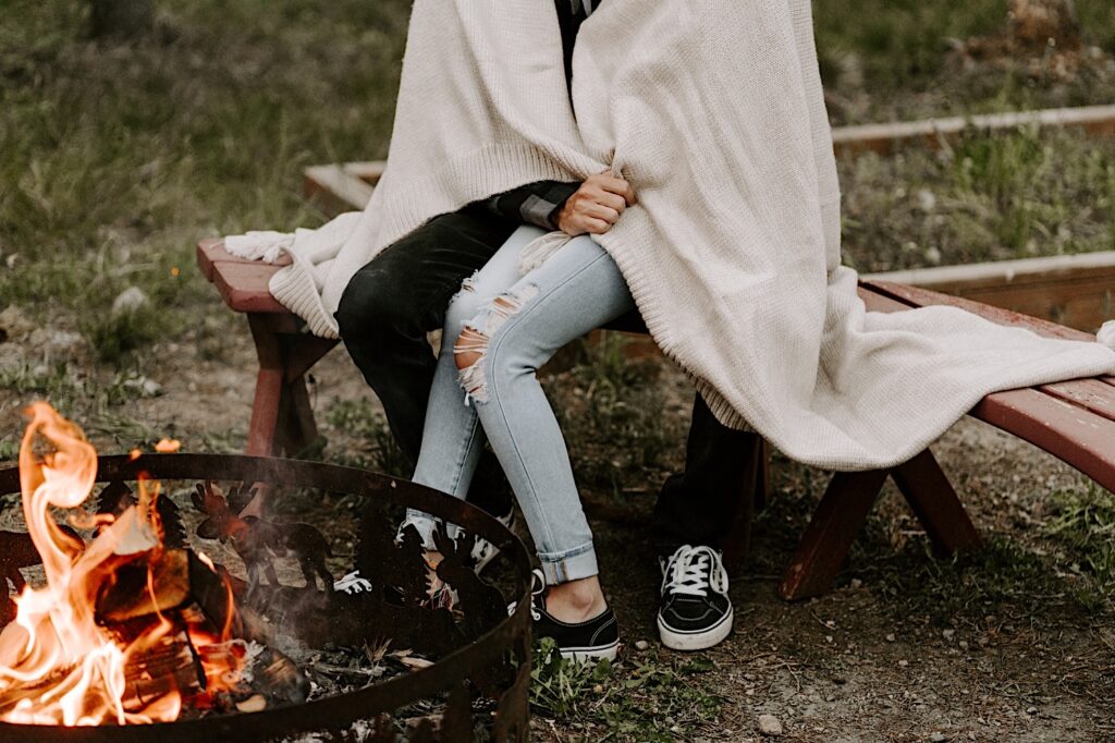 Shoulder down photo of a man and a woman wrapped under a blanket sitting on a bench in the woods next to a fire pit