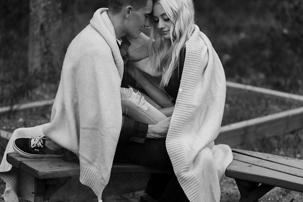 Black and white photo of a man and a woman sitting on a bench under a blanket touching foreheads together in the middle of Busse Woods Forest Preserve during their engagement session