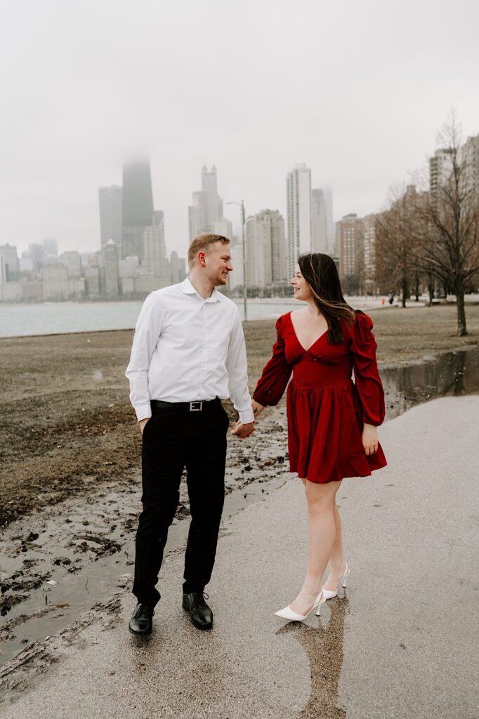 A man and woman walk along a sidewalk in a park while holding hands and smiling at one another with the Chicago skyline and Lake Michigan behind them