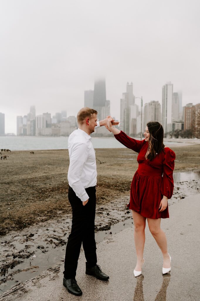 A man and woman dance together on a sidewalk in a park while smiling with Lake Michigan and the Chicago skyline behind them