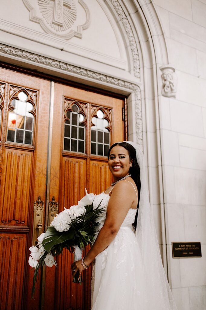 A bride in her wedding dress smiles over her shoulder at the camera while waiting outside of the entrance to a church for her wedding ceremony
