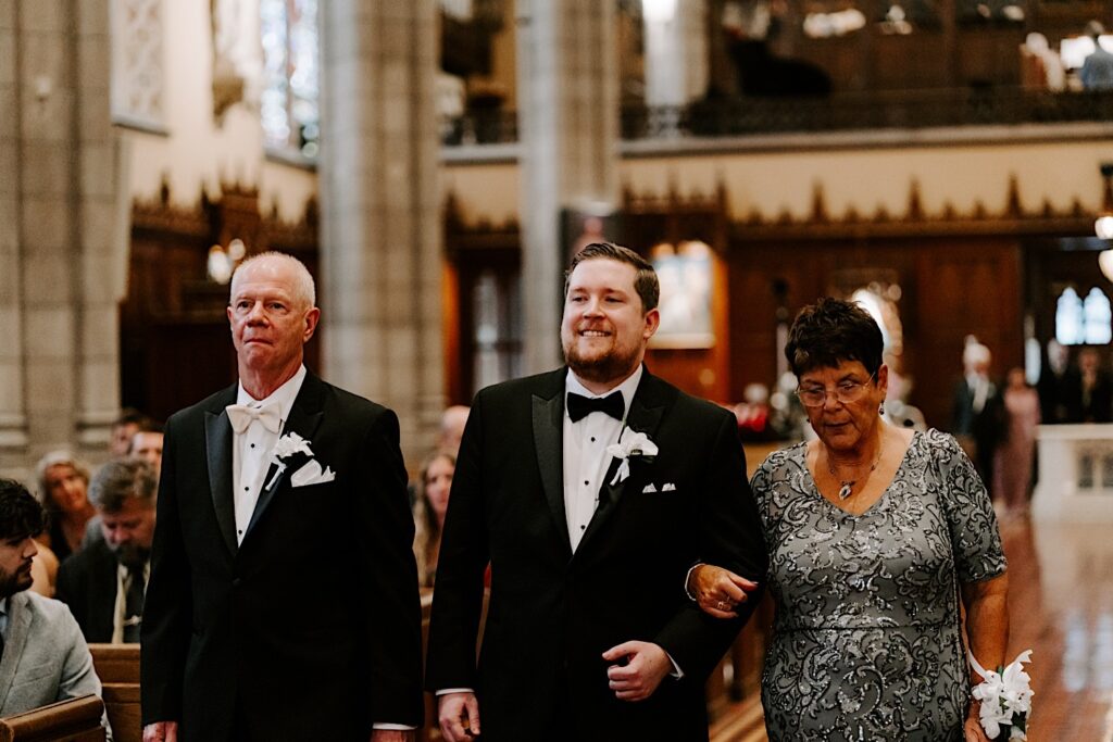 A groom smiles while walking down the aisle of a church for his wedding ceremony while being escorted by his parents