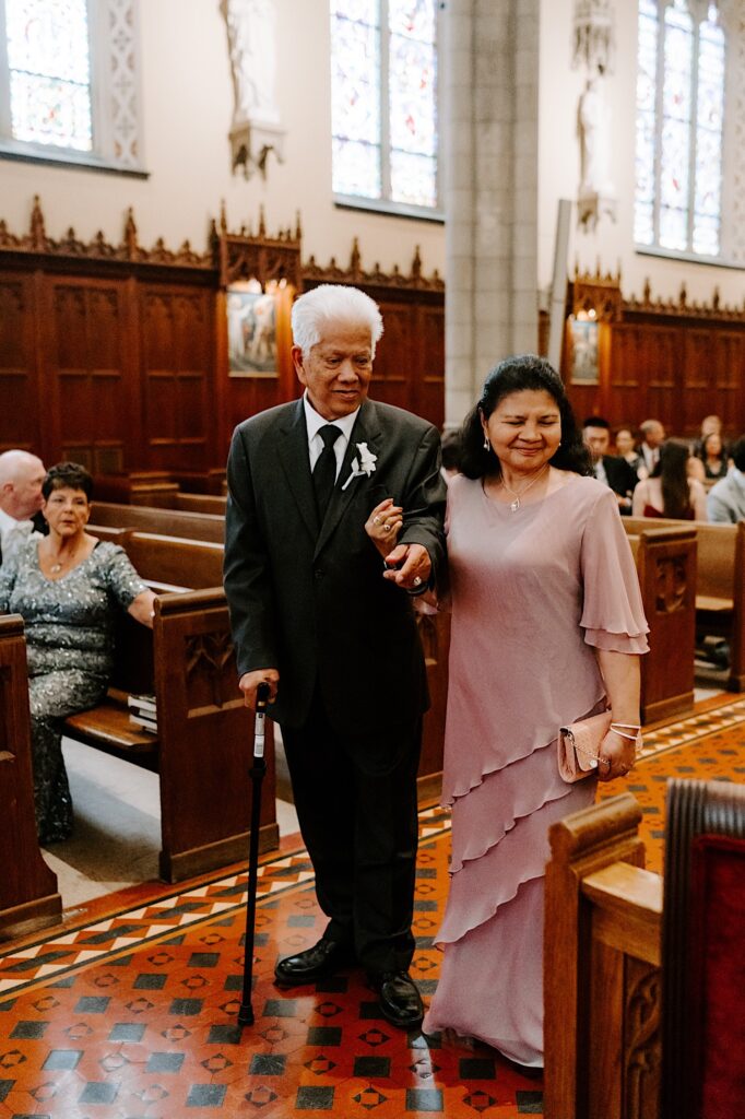 An older couple stand together and smile as they're about to sit down in on a church pew