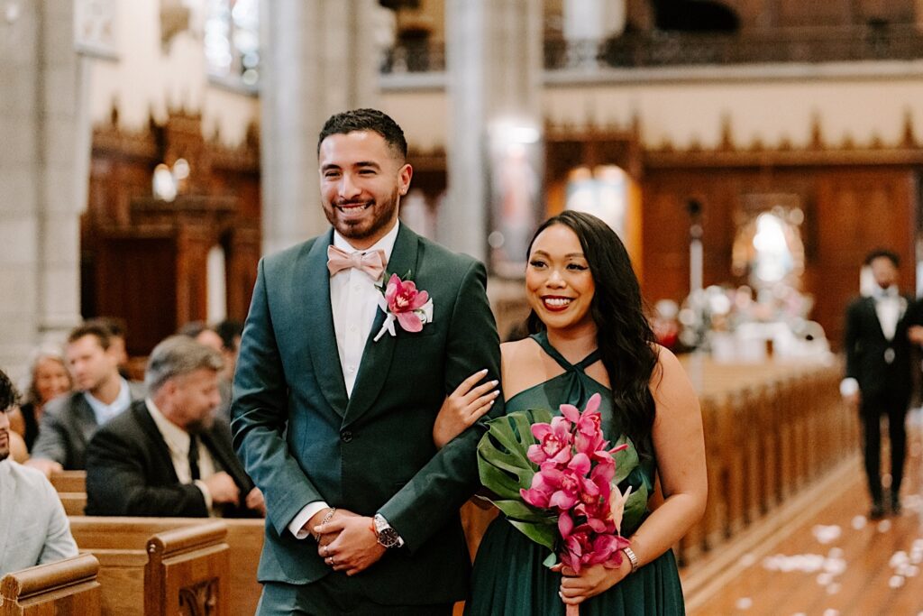 A groomsman and bridesmaid smile as they walk down the aisle together during a wedding ceremony in a church