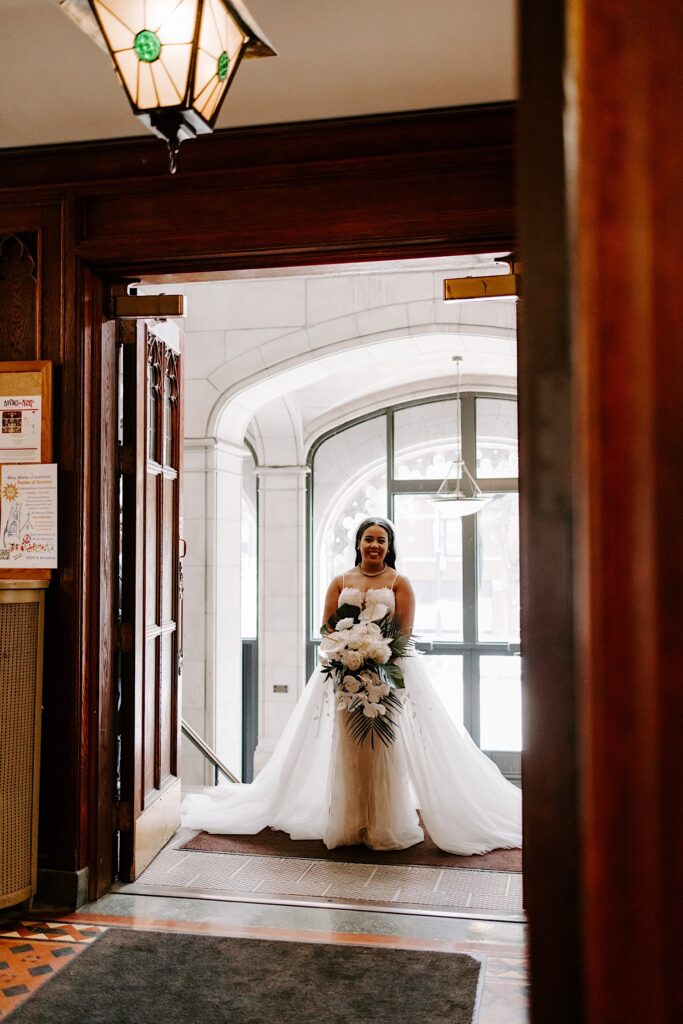 A bride smiles as she stands in an open doorway with windows behind her as she is about to enter a church for her wedding ceremony