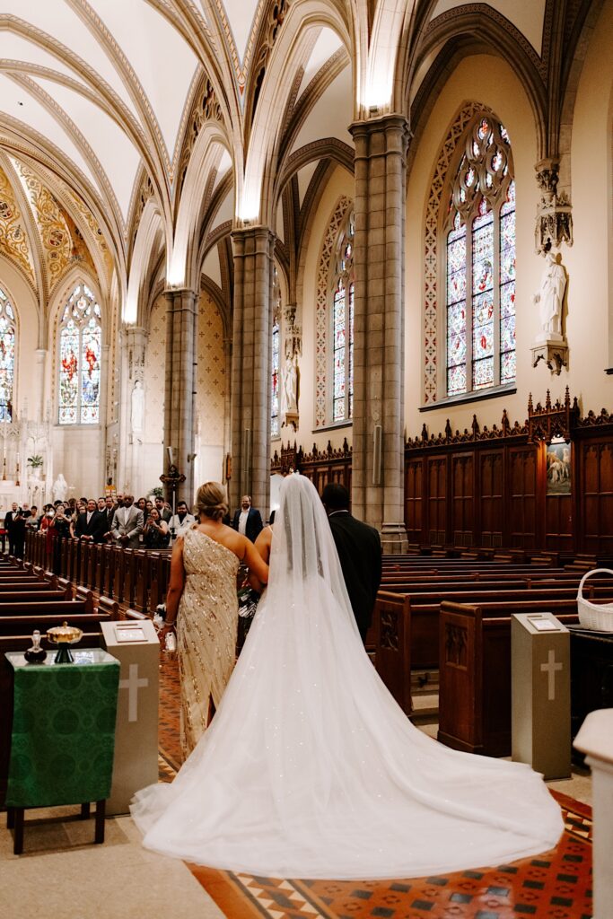 A bride walks down the aisle away from the camera while being escorted by her parents during her wedding ceremony in a church