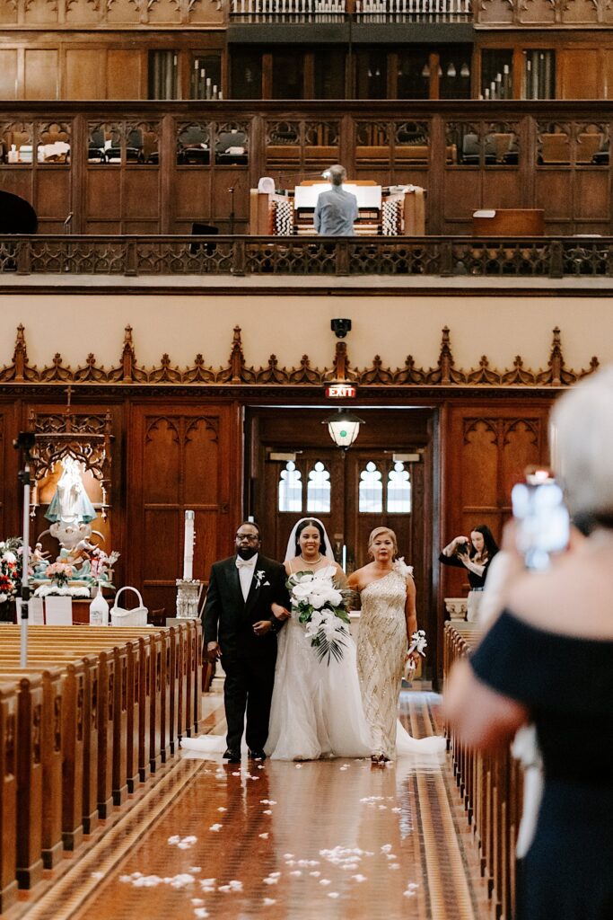 A bride smiles as her parents walk her down the aisle of a church for her wedding ceremony as guests stand and take pictures in front of her