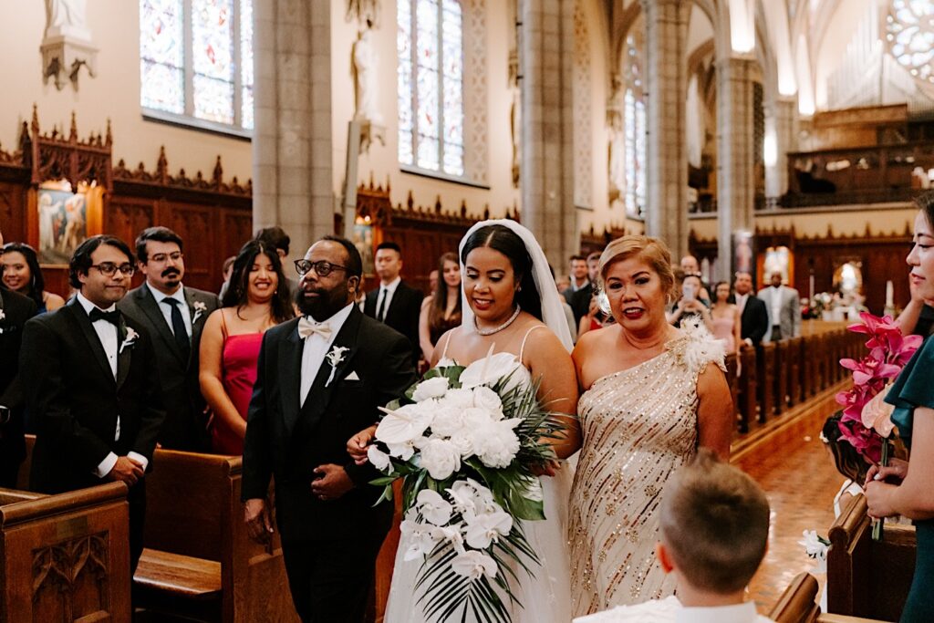A bride smiles while walking down the aisle of a church with her parents as guests on either side of them stand and watch