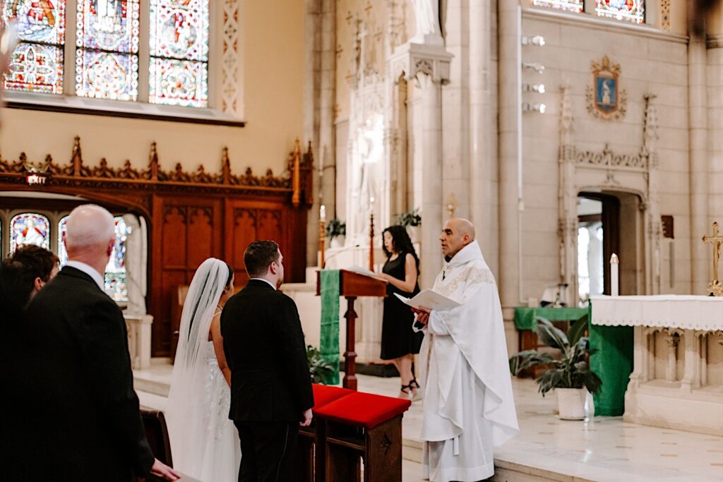 A bride and groom stand together at the altar in a church as a priest in front of them speaks during their wedding ceremony