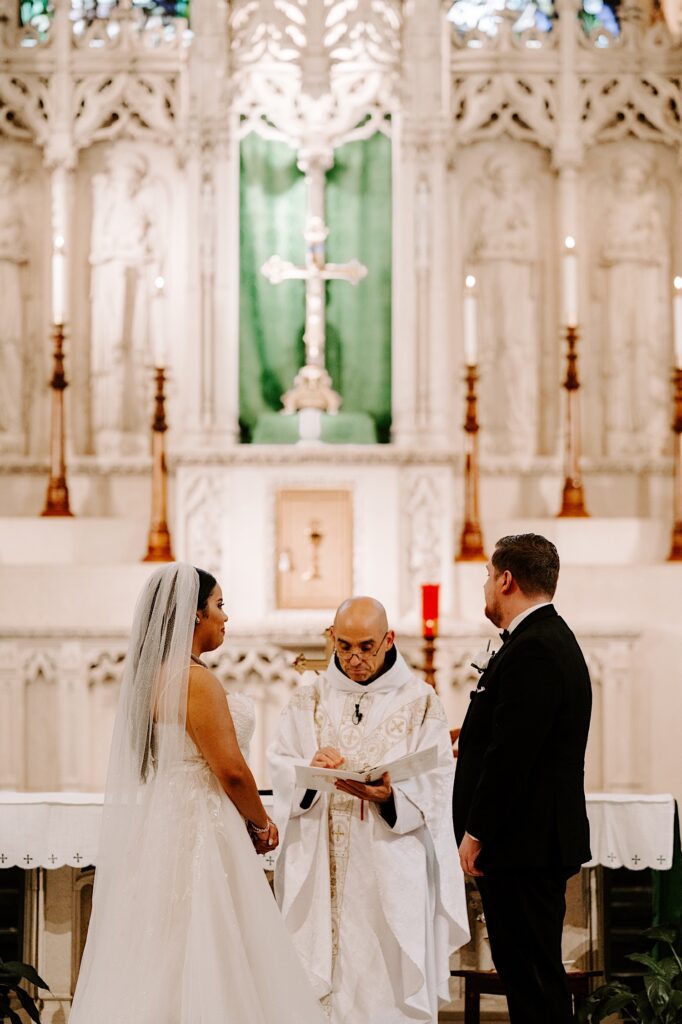 A bride and groom stand facing one another while a priest in between them speaks during their wedding ceremony inside of a church
