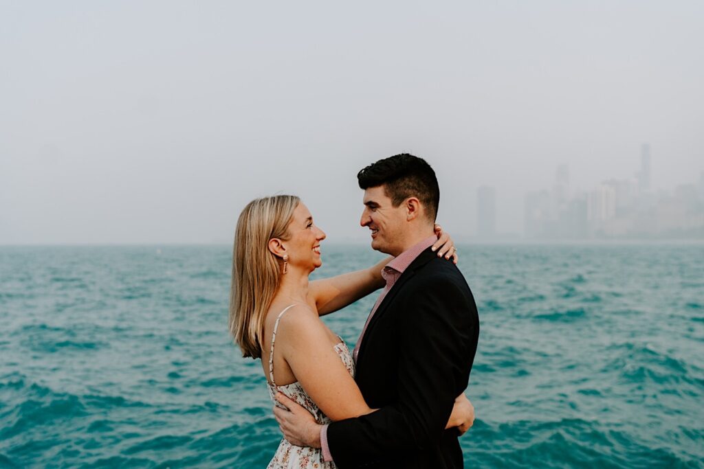 A couple embrace while smiling at one another with Lake Michigan and a hazy Chicago skyline in the background behind them