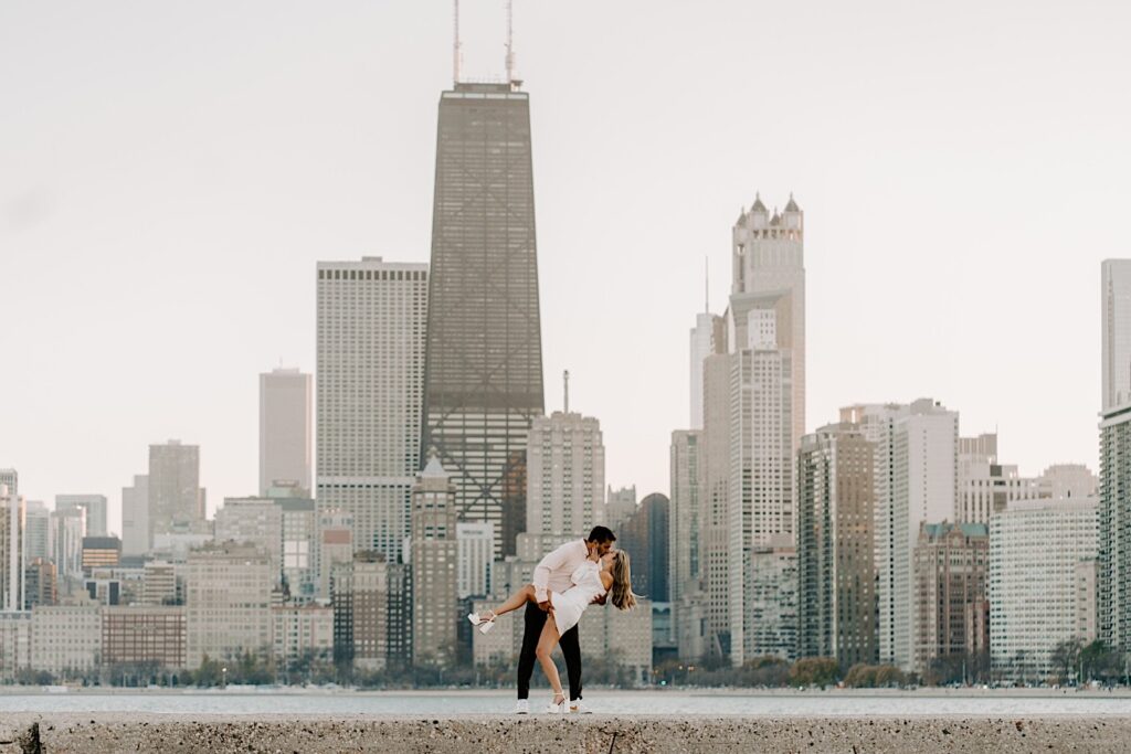 A man kisses a woman while dipping her during their engagement portraits at North Avenue Beach, behind them is the Chicago skyline