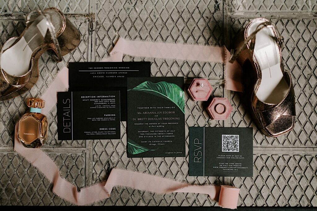 A wedding day flat lay consisting of invites, shoes, perfume, wedding rings, and a roll of fabric on a tile floor