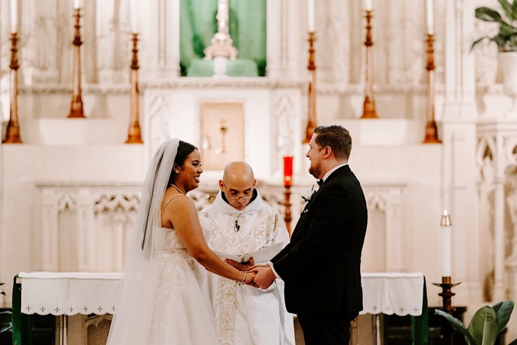 A bride and groom face each other and hold hands while smiling during their wedding ceremony as a priest in front of them speaks 