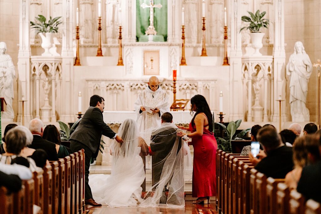 A bride and groom kneel at the altar while members of the wedding drape the bride's veil over the two of them, in front of them is the priest who is giving a speech during their ceremony in a church