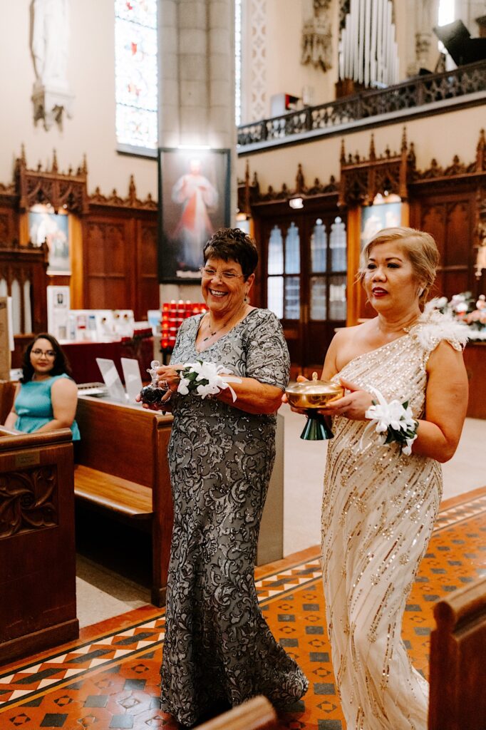 2 mothers walk down the aisle of a church together and smile as each of them carries an item towards the altar