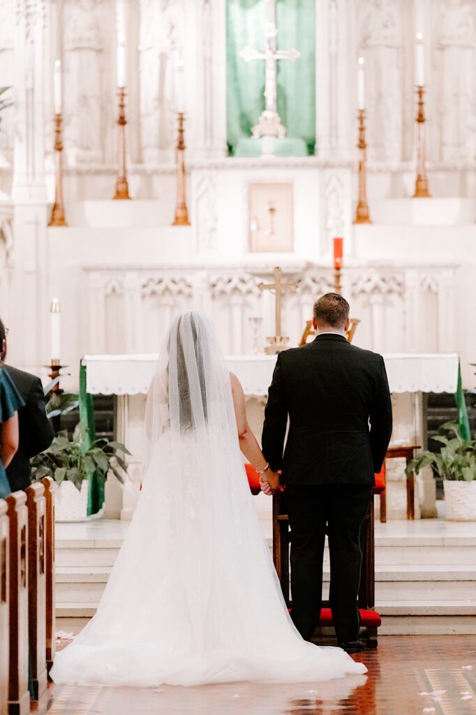 A bride and groom stand together at the altar and hold hands while facing away from the camera towards the cross of the church