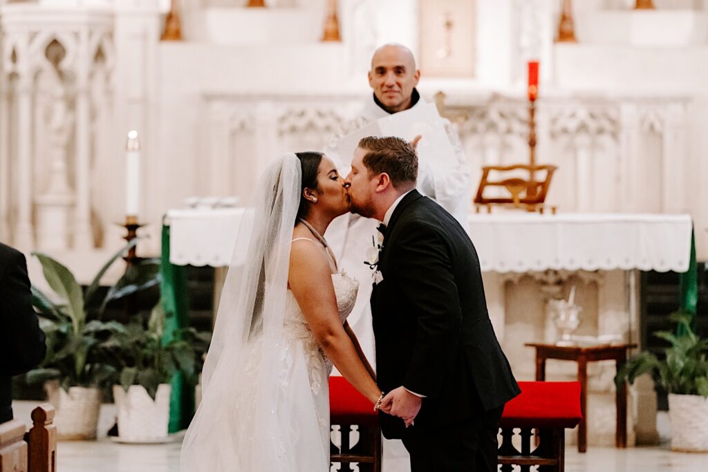 A bride and groom kiss one another while standing at the altar as the priest watches and claps during their wedding ceremony in a church