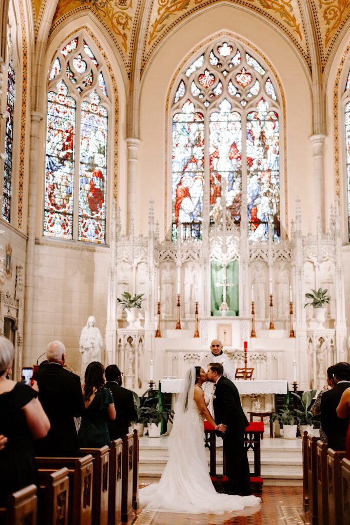 A bride and groom kiss one another while standing at the altar of a church as guests cheer for them