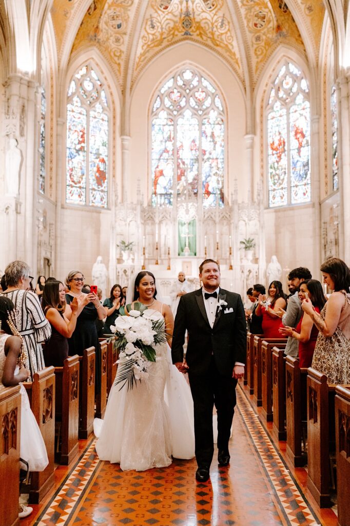 A bride and groom walk hand in hand down the aisle and smile as guests cheer for them after their wedding ceremony in a church