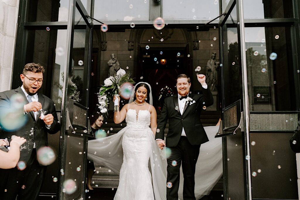 A bride and groom exit a church and smile while holding hands as bubbles fill the air around them, blown by guests of the wedding standing outside