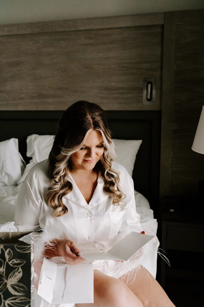 A bride sits on a bed before getting dressed for her wedding, as she sits on the bed she smiles while reading a letter