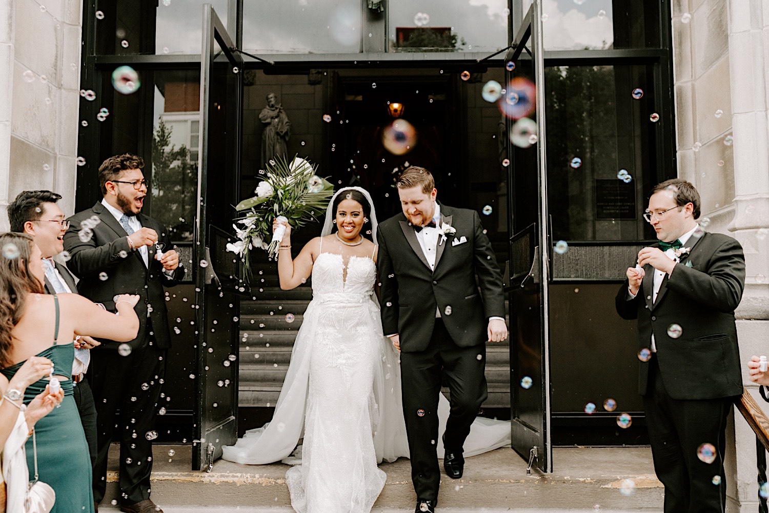 Guests of a wedding cheer and blow bubbles as the bride and groom exit the church and smile while holding hands after their wedding ceremony