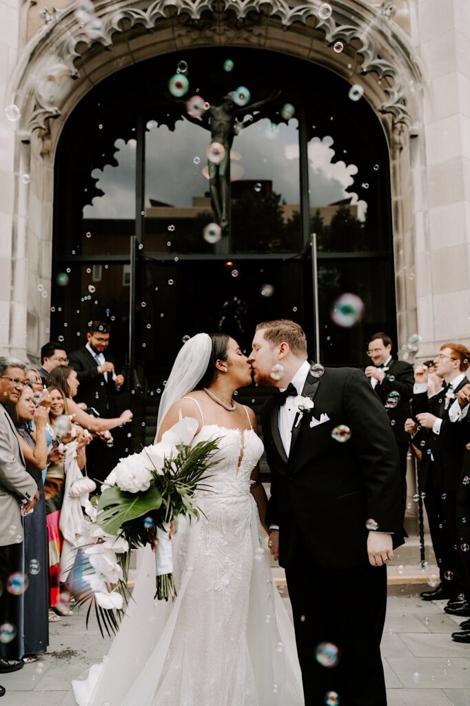 A bride and groom kiss while outside a church as guests of their wedding surround them and blow bubbles