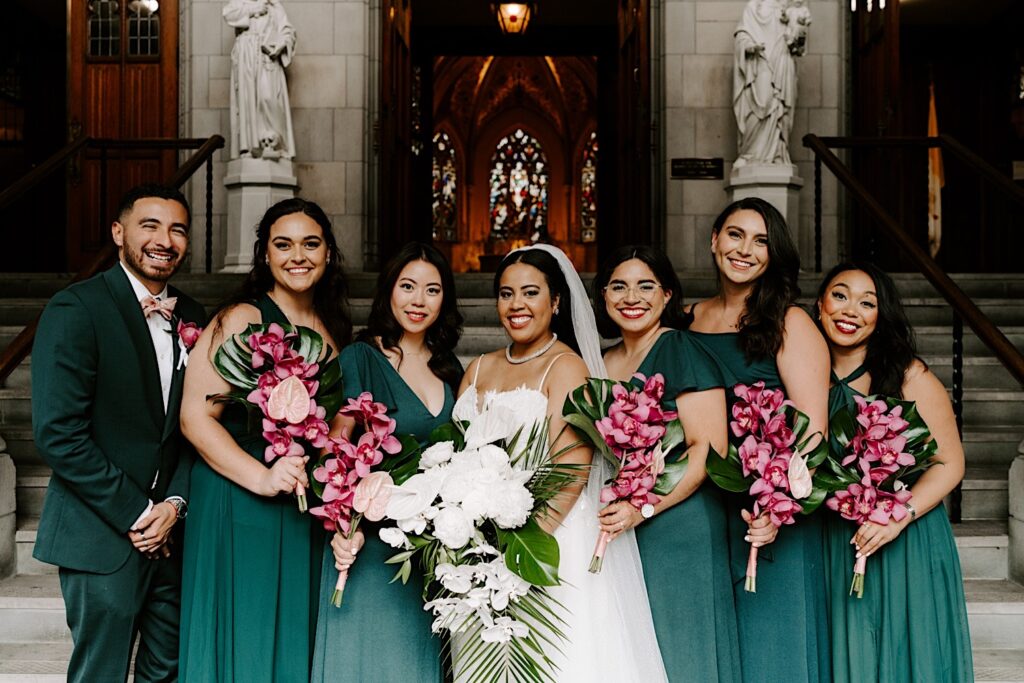A bride stands with the 6 members of her wedding party in front of a church as they all smile at the camera
