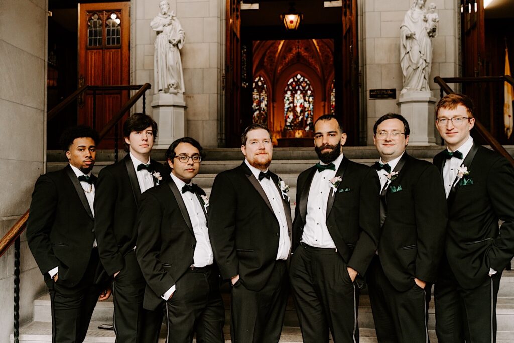 A groom stands with the 6 members of his wedding party in front of a church as they all pose with their hands in their pockets