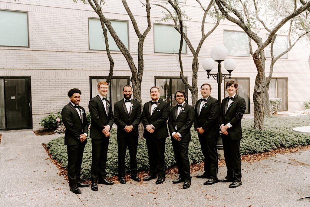 A groom stands with his 6 groomsmen in a courtyard as they all smile at the camera while clasping their hands