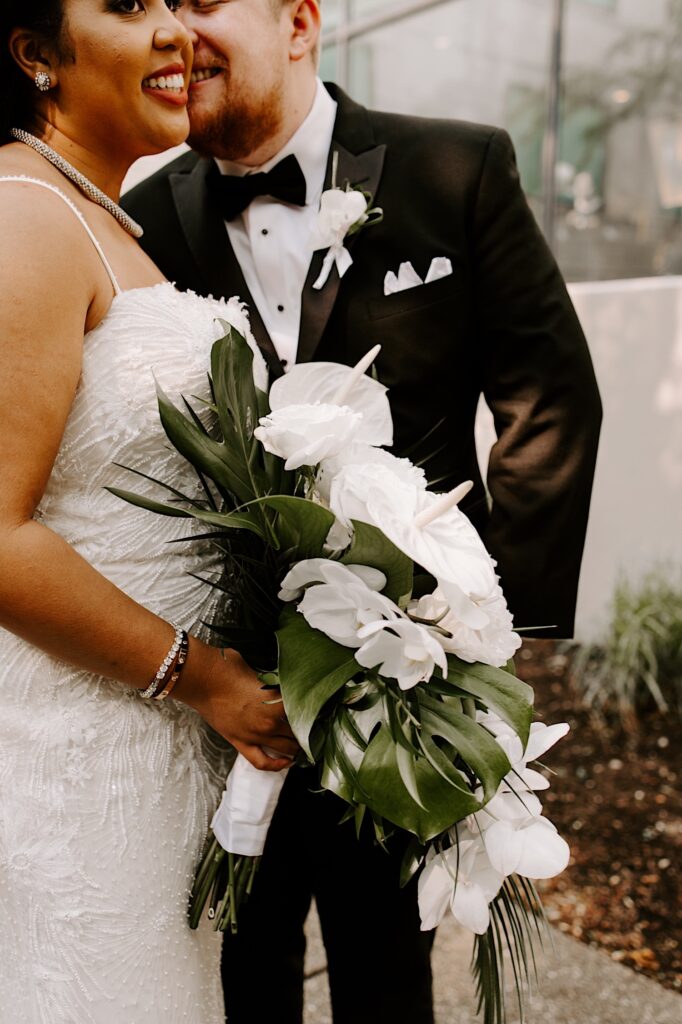 A bride smiles while holding her white floral bouquet as the groom is about to kiss her on the cheek while the two stand outside